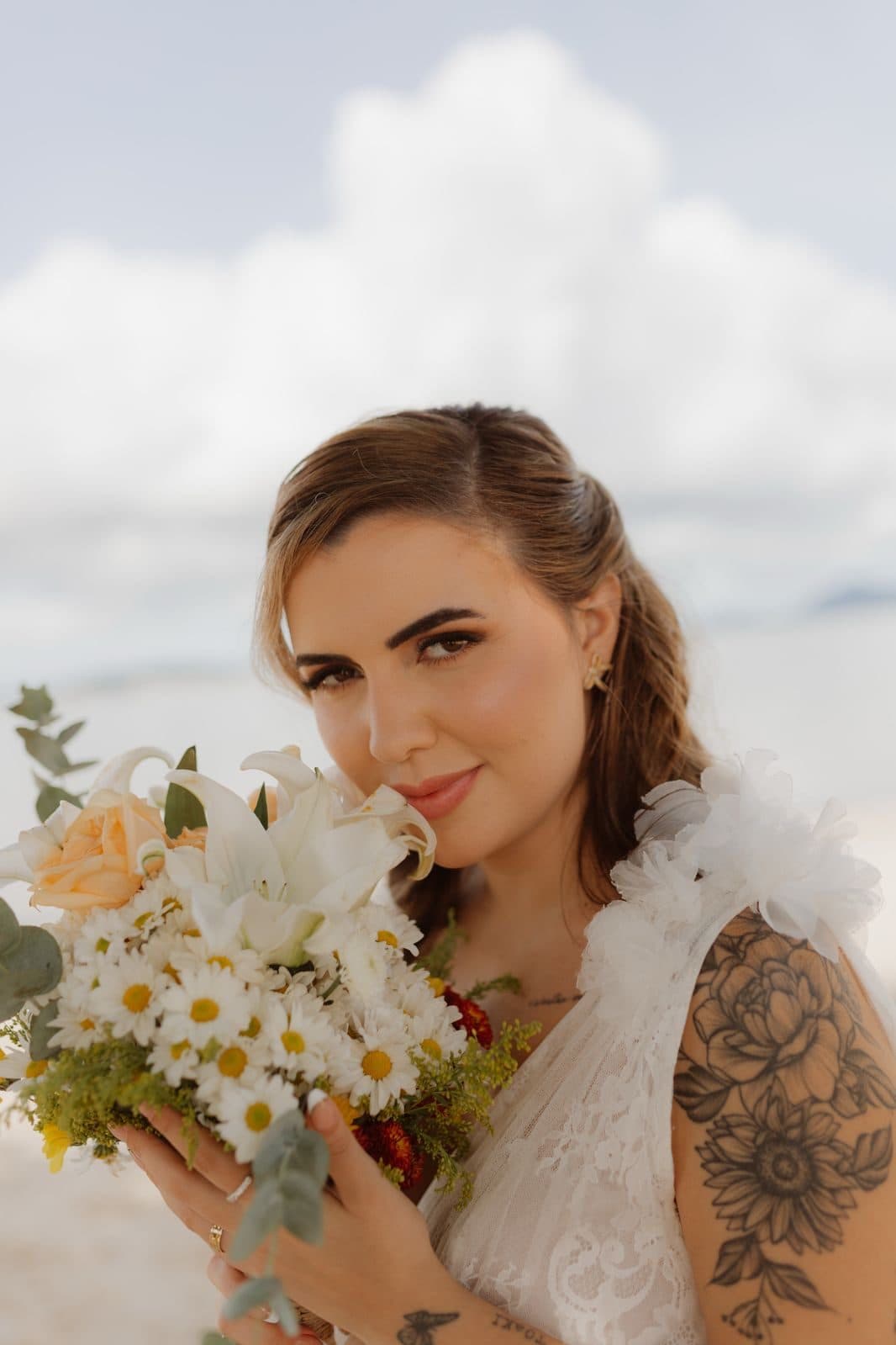 Noiva com tatuagem no braço segurando buquê de margaridas contra o céu