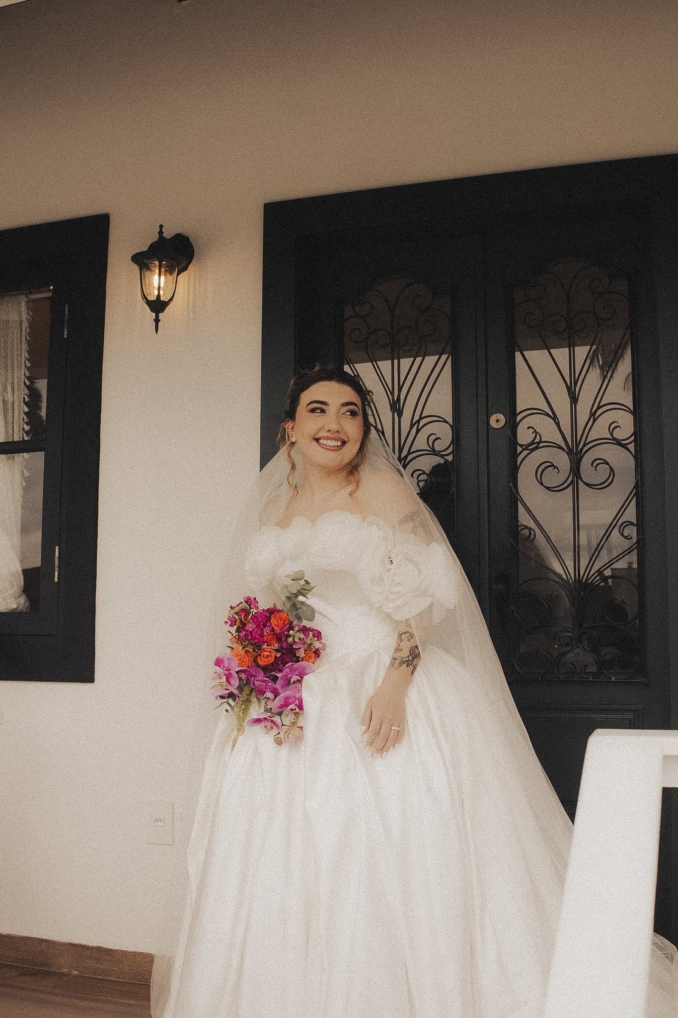 Noiva sorrindo na varanda, com véu longo e buquê de orquídeas e rosas