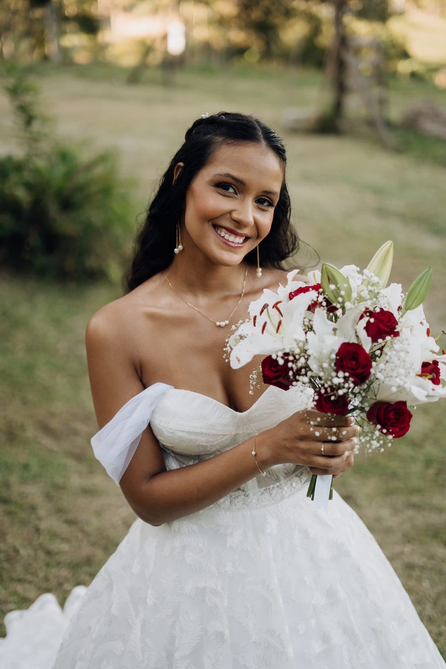 Noiva sorrindo ao ar livre com buquê de rosas vermelhas e lírios