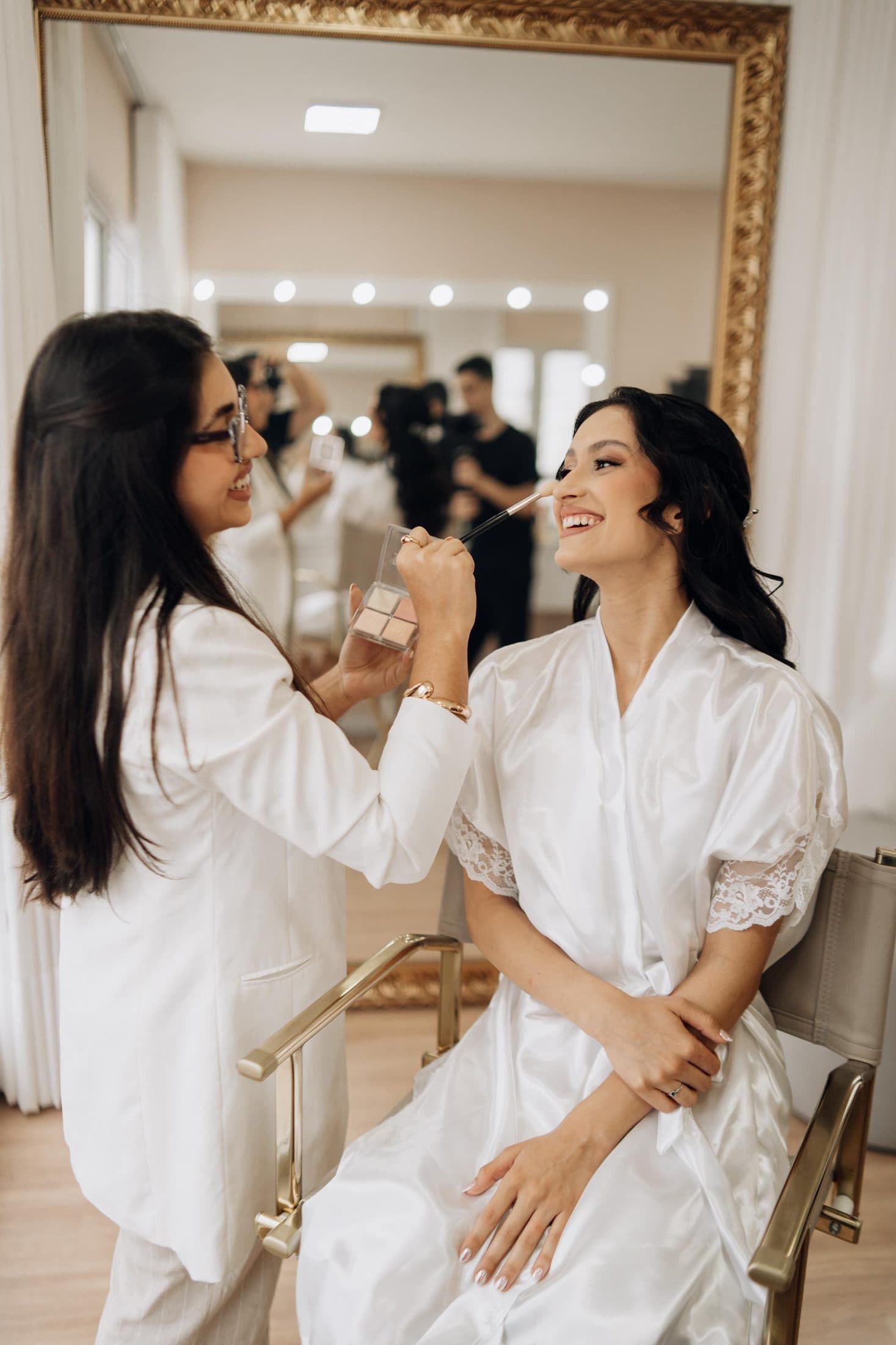 Dani Rabello maquiando uma noiva de robe branco em frente a um espelho de moldura dourada