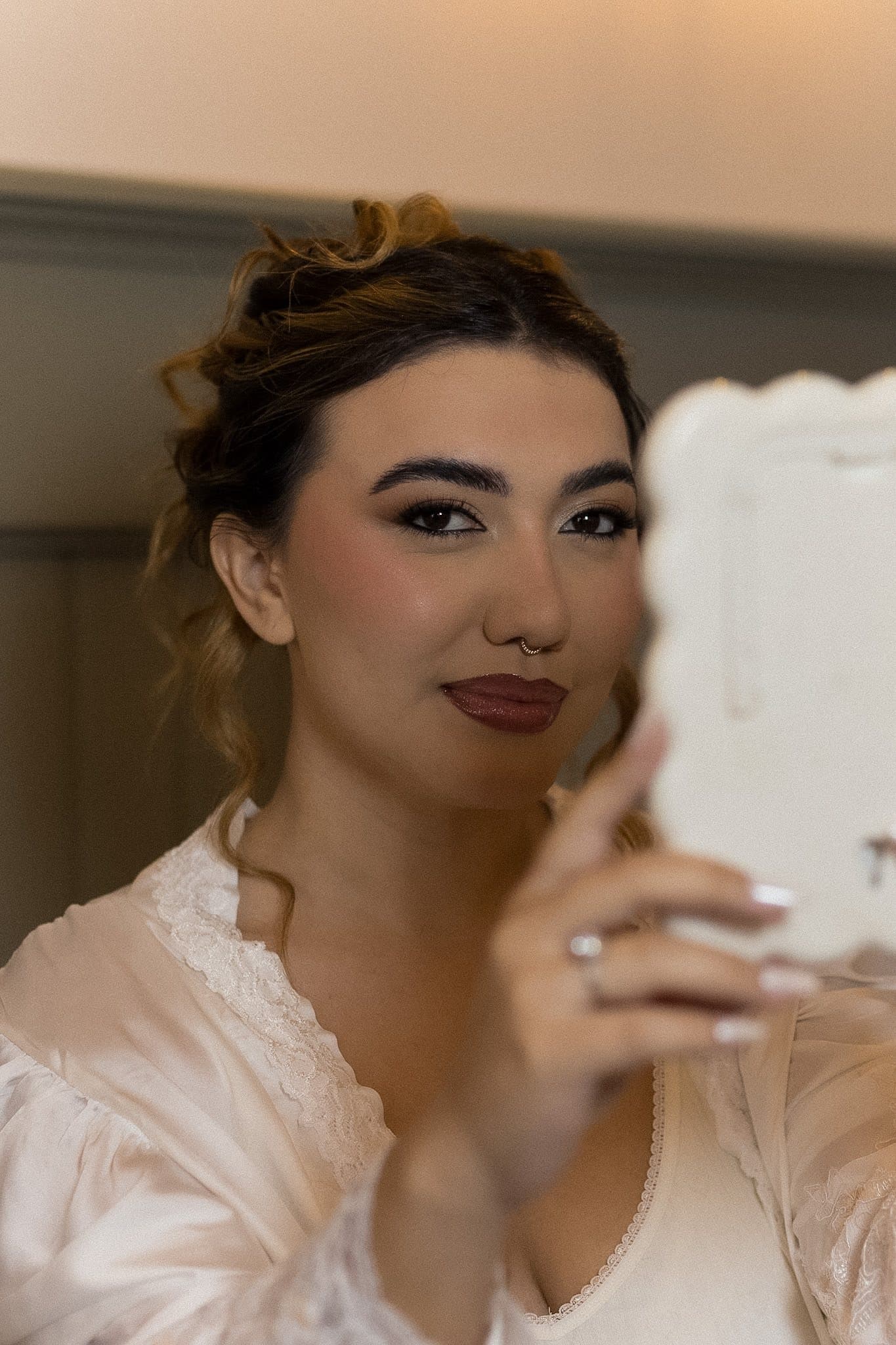 Noiva de robe branco se olhando no espelho de mão, com coque baixo e batom vinho