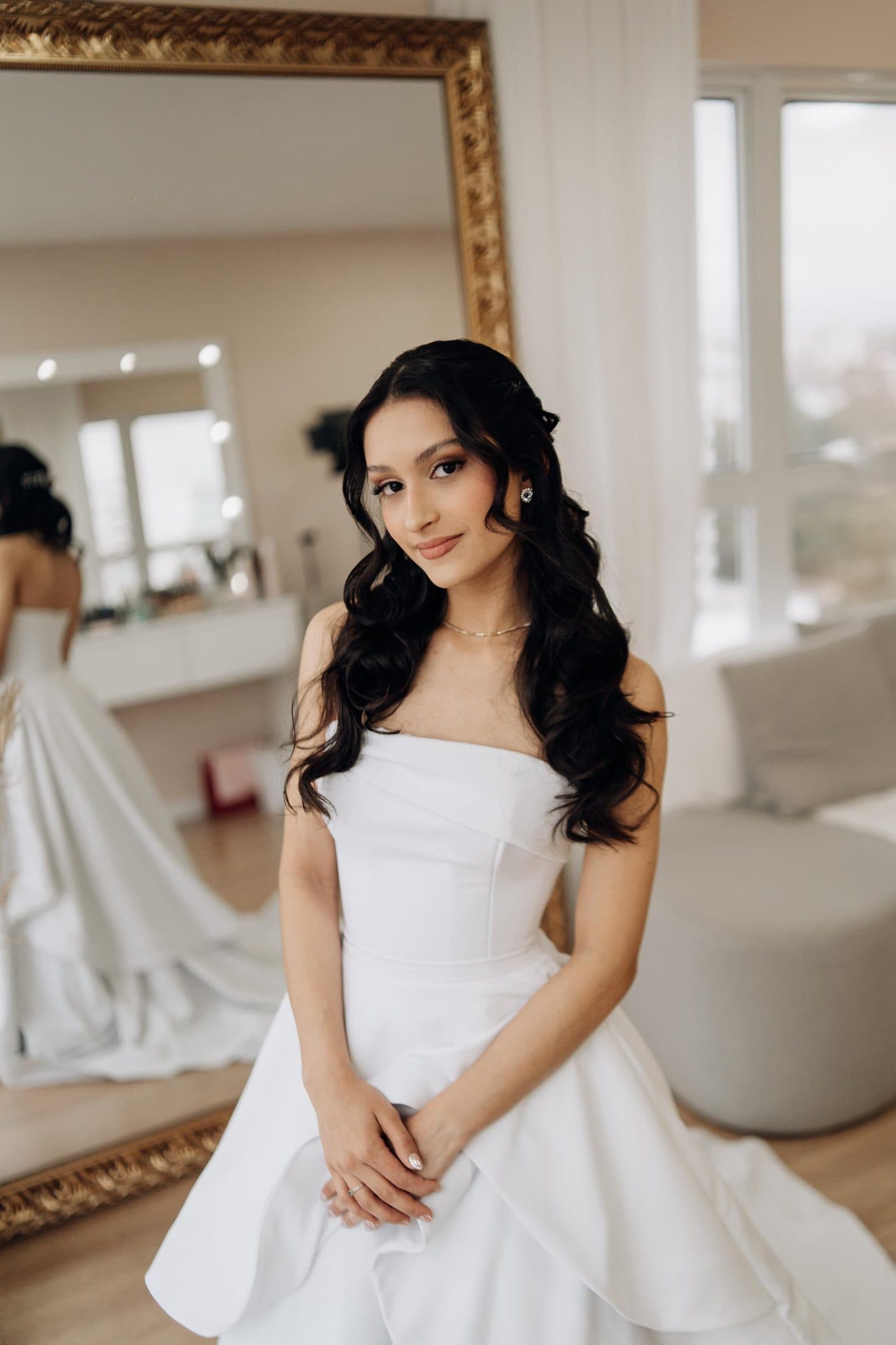 Noiva de cabelo solto e ondulado sentada em uma suíte clara, já pronta
